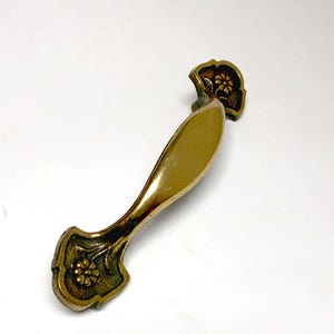 May include: A brass-toned metal drawer pull featuring a smooth, elongated central handle and two flared ends. Each end is decorated with a small, embossed floral motif set against a textured, darkened background, providing a contrast to the polished finish.