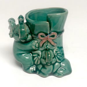 May include: A teal ceramic planter shaped like a boot with two sculpted elephants attached to the front. The boot features a small pink bow detail on the side. The glazed ceramic surface has a visible crackle finish throughout the entire piece.