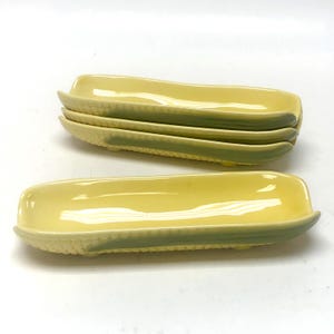 May include: A set of four elongated ceramic corn-on-the-cob dishes. Each dish features a glossy yellow interior and a textured exterior resembling corn kernels, with a pale green base. The dishes are arranged with three stacked and one in front.