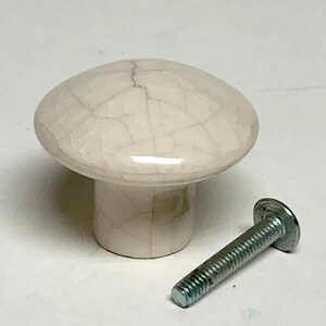 May include: A round, off-white ceramic cabinet knob featuring a glossy finish with a dark, web-like crackle pattern across the surface. The knob has a cylindrical base and is accompanied by a separate silver-colored metal bolt with a flat head.