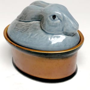 May include: A ceramic lidded dish featuring a sculpted, glossy light blue rabbit figurine as the lid handle. The base of the dish is oval-shaped with a smooth, matte terracotta-colored exterior and a dark, charcoal-colored rim at the top edge.