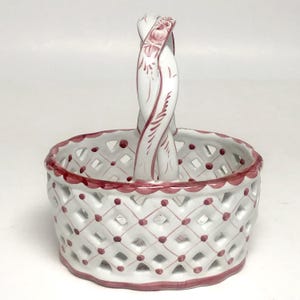 May include: A white ceramic basket with a lattice design and a single arched handle. The basket features hand-painted pink accents along the rim, the handle, and at each intersection of the diamond-shaped lattice pattern, creating a decorative look.