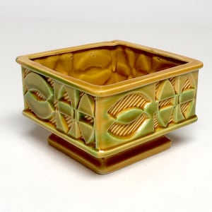 May include: A square ceramic planter with a glossy finish in shades of mustard yellow and sage green. The exterior features a repeating geometric pattern of embossed leaf-like shapes and diagonal lines, set on a raised square base.