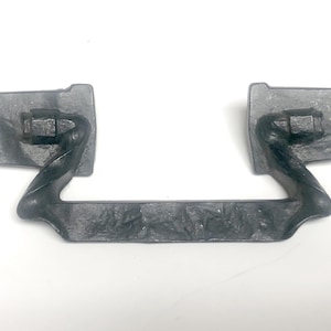 May include: A dark gray metal drop handle featuring a textured, hammered finish. The hardware consists of two rectangular mounting plates connected by a central, swinging pull bar with a twisted design on each side and a flat, textured center.