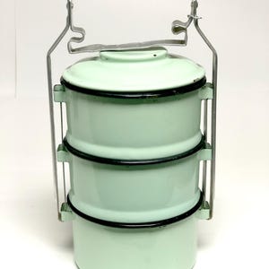 May include: A vintage, three-tiered, mint green enamel food carrier with black trim. It features a metal handle with a black wooden grip and a metal carrying frame. The carrier is designed to stack and transport food items.