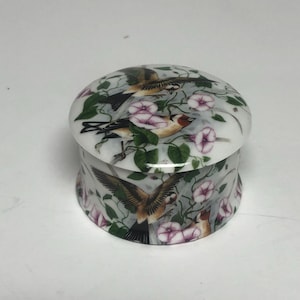 May include: A small, round trinket box with a lid. The box is white with a detailed illustration of birds, pink flowers, and green leaves. The birds have brown, black, and yellow feathers. The box is likely made of ceramic or porcelain.