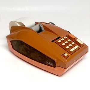 May include: A vintage Sears electric adding machine with a brown plastic casing and a wood-grain side panel. The device features a numeric keypad with off-white buttons, a paper roll holder at the back, and a small red switch on the top surface.