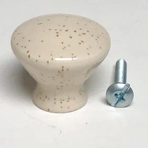 May include: A round, cream-colored cabinet knob with a glossy finish and brown speckles throughout the surface. The knob features a flared base and is accompanied by a single silver-toned metal machine screw with a Phillips head for installation.