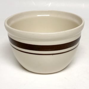 May include: A cream-colored ceramic mixing bowl featuring a wide dark brown horizontal band encircling the exterior, complemented by a thinner dark brown line positioned directly below the wider band, creating a simple and functional kitchen vessel.