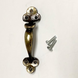 May include: A brass-toned metal cabinet handle with a dark, weathered finish and a curved, elongated center. The handle features decorative, clover-shaped ends with small circular indentations. Two silver-colored metal screws are included for installation.