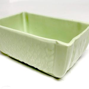 May include: A rectangular light green ceramic planter featuring a textured exterior with a repeating leaf-like pattern. The vessel has a smooth interior, a flat base, and slightly flared corners, providing a simple and functional container design.