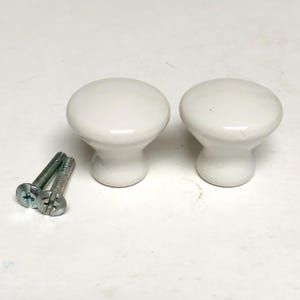 May include: Two smooth, glossy white ceramic cabinet knobs with a rounded top and a tapered base. Each knob features a circular face and a narrow neck. Two silver-colored metal machine screws with flat heads are positioned next to the knobs.