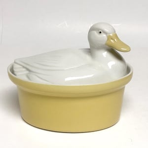 May include: A ceramic lidded casserole dish featuring a white duck-shaped lid with a light yellow beak. The base of the dish is a solid, smooth light yellow color with a rounded, cylindrical shape. The duck lid has molded feather details.
