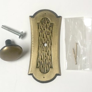 May include: A brass-toned metal door plate with a textured center panel, a matching round knob, a long mounting screw, and two small nails in a clear plastic bag. The rectangular plate features a central hole and a decorative finish.