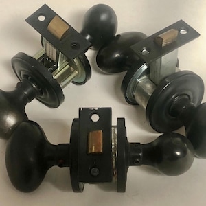 May include: A collection of black door knobs and latches. The knobs have an oval shape and are made of a dark, possibly metal material. The latches have a rectangular plate with a brass-colored piece. The hardware is likely for interior doors.