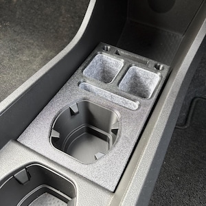 May include: A custom-fit, textured black car console insert with cup holders and storage compartments. The insert is designed to fit snugly within the vehicle's existing console, providing additional organization and functionality. The design includes two cup holders and two storage compartments.