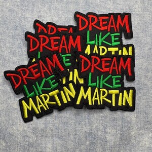 May include: Embroidered patches with the text "DREAM LIKE MARTIN" in red, green, and yellow lettering on a black background. The patches are stacked on a light blue fabric surface.