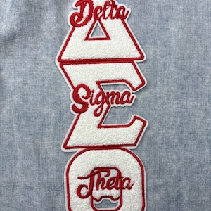 May include: A white and red embroidered patch featuring the Greek letters Delta, Sigma, and Theta. The letters are in a stacked arrangement, with red script lettering on a white, textured background. The patch is likely for a sorority or fraternity.