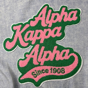 May include: A green and pink embroidered patch with the words "Alpha Kappa Alpha" in a cursive font. The patch also includes the text "Since 1908" in green. The patch is on a light blue fabric.