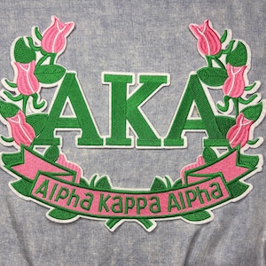 May include: Embroidered patch featuring the letters "AKA" in green, surrounded by a wreath of pink roses and green leaves. A pink banner with the words "Alpha Kappa Alpha" is below the letters. The patch is on a gray fabric background.