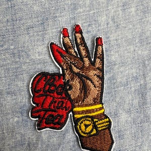 May include: Embroidered patch featuring a brown hand with red fingernails and a gold watch. The patch includes the text "Cheek The Tea" in red and white, with a white border. The patch is on a blue denim background.