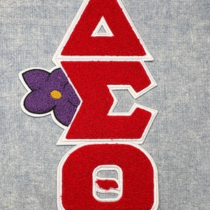 May include: A red and white embroidered patch featuring the Greek letters Delta, Sigma, and Omega, with a purple flower accent. The patch is on a light blue denim background. The letters are outlined in white.