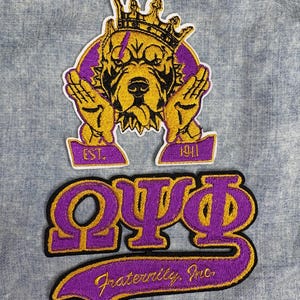 May include: Two embroidered patches. The top patch features a crowned dog with outstretched hands, in gold and purple. The bottom patch displays Greek letters and the words "Fraternity, Inc." in purple and gold. The patches are on a light blue denim background.
