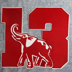 May include: A red felt patch featuring the number "13" with a white outline. An elephant graphic is centered on the patch. The elephant is red with white accents. The patch is likely for a sports team or school.