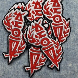 May include: A collection of red and white embroidered patches with a black border. Each patch features the letters "AHO" in a stylized font, surrounded by a laurel wreath design. The patches are on a light blue denim background.
