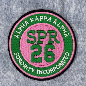 May include: Embroidered patch with a circular design. The patch features the text "ALPHA KAPPA ALPHA SORORITY INCORPORATED" in white, with "SPR 26" in green. The background is pink and green, with a black border.