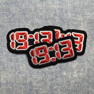 May include: A cluster of four black embroidered patches with the numbers "19:13" in red, outlined in white. The patches have a black border and are arranged on a light blue textured fabric.