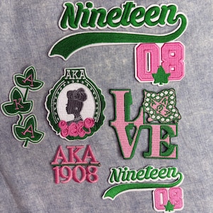 May include: A collection of embroidered patches in green and pink, featuring the words "Nineteen 08" and "LOVE". Other patches include the letters "AKA" and a silhouette of a woman. The patches are on a light blue fabric.