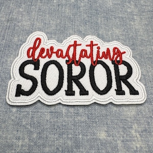May include: Embroidered patch with the words "devastating SOROR" in red and black thread. The text is on a white background with a scalloped edge. The patch is on a blue denim background.