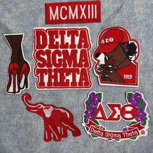May include: A collection of embroidered patches in red and white, featuring the text "DELTA SIGMA THETA", an elephant, a woman in a red cap, and the Roman numeral "MCMXIII". The patches are on a light blue fabric.