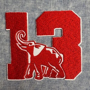 May include: A red embroidered patch featuring the number "13" with a white-outlined elephant graphic. The patch is on a light blue denim-like fabric. The elephant is in the center of the number.