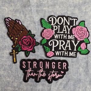 May include: Three embroidered patches on a blue fabric background. One patch shows praying hands with a cross and a pink rose. Another says "DON'T PLAY WITH ME PRAY WITH ME" with pink roses. The third says "STRONGER than the storm".