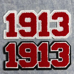 May include: Two embroidered patches with the number "1913". The top patch has red numbers with a white border, while the bottom patch has red numbers with a black border. Both patches are on a light blue fabric.