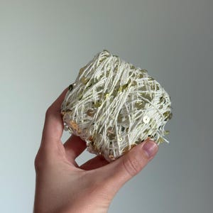 May include: A ball of off-white yarn featuring small, reflective gold-colored sequins distributed throughout the fibers. The yarn is wound into a compact, rounded shape, showcasing the contrast between the matte thread and the metallic embellishments.