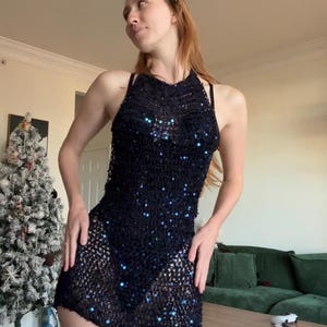 May include: A black halter dress with a mesh design and sparkling sequins. The dress is sleeveless and has a mini length. The dress is worn over a black undergarment. The dress is a part of the clothing category.