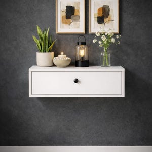 以下が含まれることがあります： 黒いノブが付いた白いフローティングドロワーが、濃い灰色の壁に取り付けられています。 棚には、鉢植えの植物、ろうそく、装飾的なボウル、ランタン、花瓶の花、2つの額入りの抽象芸術作品が飾られています。