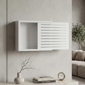 May include: A white wall-mounted cabinet with a sliding door featuring horizontal slats. The cabinet is open, revealing an empty interior. The image also shows a vase with branches, books, and a gold clock.