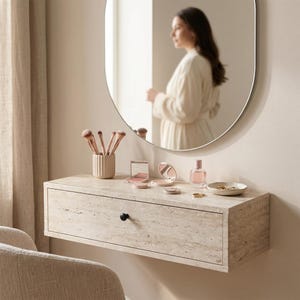 以下が含まれることがあります： 引き出し付きのフローティングバニティ。メイクアップとブラシが入っています。丸い鏡が上にあり、白いローブを着た人を映しています。バニティは明るい色の石でできており、引き出しには黒いノブが付いています。
