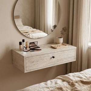 Puede incluir: Un tocador flotante con acabado tipo mármol y tirador negro. Un espejo redondo refleja una cama. Productos de maquillaje y una bandeja de madera con objetos pequeños sobre la superficie. Predominan los tonos neutros.
