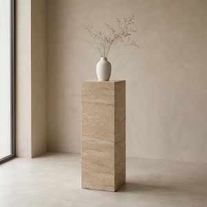 May include: A rectangular stone pedestal in shades of beige and brown, with a white vase holding dried stems on top. The pedestal is a display stand. The scene is lit by natural light, creating a minimalist look.