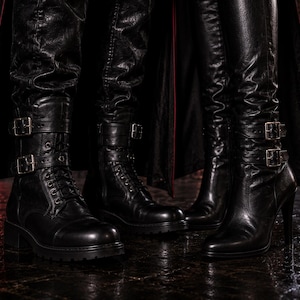 May include: Three pairs of black leather boots with buckles. The boots vary in style, including combat boots with laces and block heels, and knee-high boots with stiletto heels. The boots are on a dark, reflective surface.