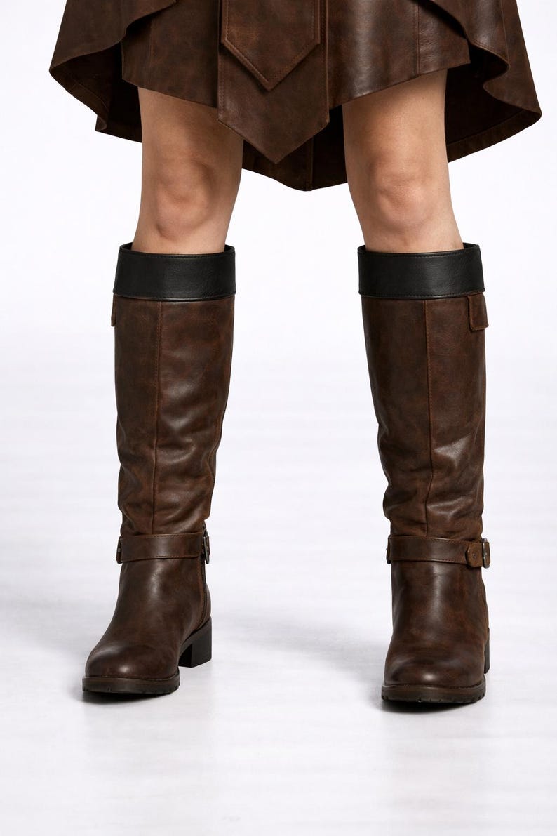 May include: Brown knee-high boots with black leather tops and buckle details. The boots have a slight heel and are made of a textured material. The boots are worn with a brown skirt.