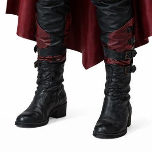 K&ouml;nnte beinhalten: Schwarze Lederstiefel mit Blockabsatz. Die Stiefel haben einen dekorativen Riemen mit Schnallen und einen roten Lederakzent. Die Stiefel werden mit einer schwarzen Lederhose und einem roten Umhang kombiniert.