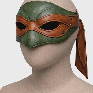 Può includere: Una maschera in pelle verde e marrone, che ricorda un personaggio popolare. La maschera presenta una sezione superiore verde e una copertura per gli occhi marrone, con un laccio abbinato sul lato. La maschera è esposta su una testa di manichino.
