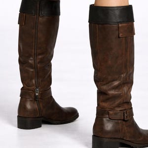 May include: Brown leather knee-high boots with a black leather top. The boots have a side zipper, a buckle strap around the ankle, and a small square heel. The boots are worn by a person.