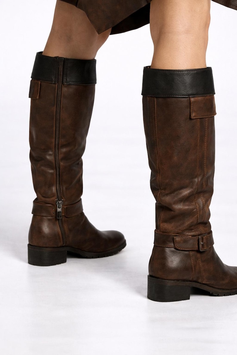 May include: Brown leather knee-high boots with a black leather top. The boots have a side zipper, a buckle strap around the ankle, and a small square heel. The boots are worn by a person.